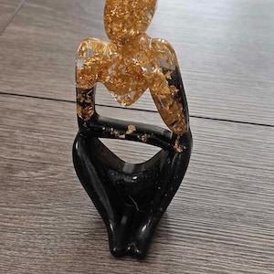 May include: A black and gold resin sculpture of a person sitting with their legs crossed. The sculpture is made of clear resin with gold leaf flakes embedded in it.