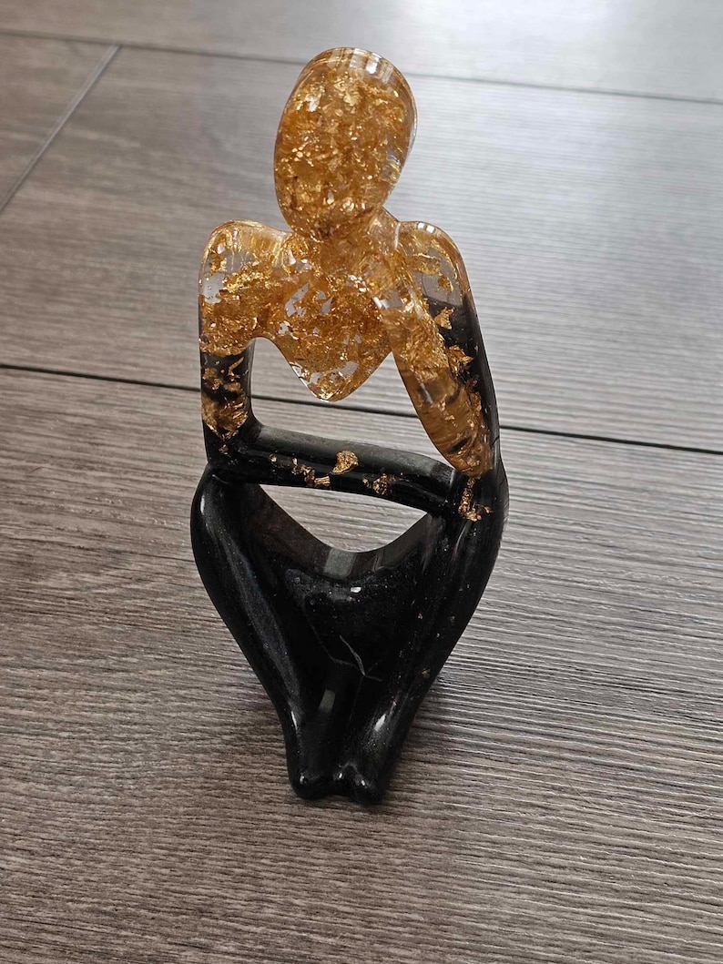 May include: A black and gold resin sculpture of a person sitting with their legs crossed. The sculpture is made of clear resin with gold leaf flakes embedded in it.