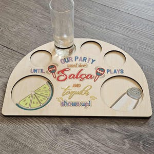Op de afbeelding: Houten dienblad voor shotglaasjes en hapjes met een halfrond ontwerp. De lade heeft uitgesneden cirkels voor shotglaasjes en kleine kommen. De tekst op de lade luidt: "OUR PARTY won't start until Salsa and Tequila shows up!" met illustraties van limoen en zoutstrooier.