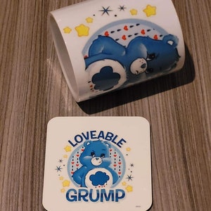 Puede incluir: Una taza de cerámica blanca y un posavasos cuadrado con una ilustración de oso azul. La taza tiene un diseño de oso durmiendo con estrellas y corazones. El posavasos dice "Loveable Grump" con una ilustración de oso gruñón.