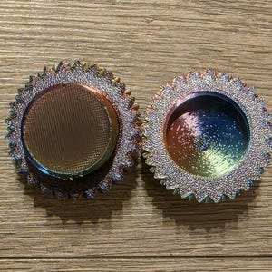 May include: Two iridescent, circular objects with a textured, ridged surface and a central, concave area. The objects have a metallic sheen with colors shifting from blue to green to purple. The outer edges are lined with small, pointed details.