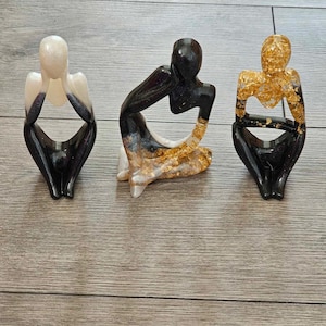 May include: Three abstract resin sculptures of people in a seated pose. The sculptures are painted in black, white, and gold.