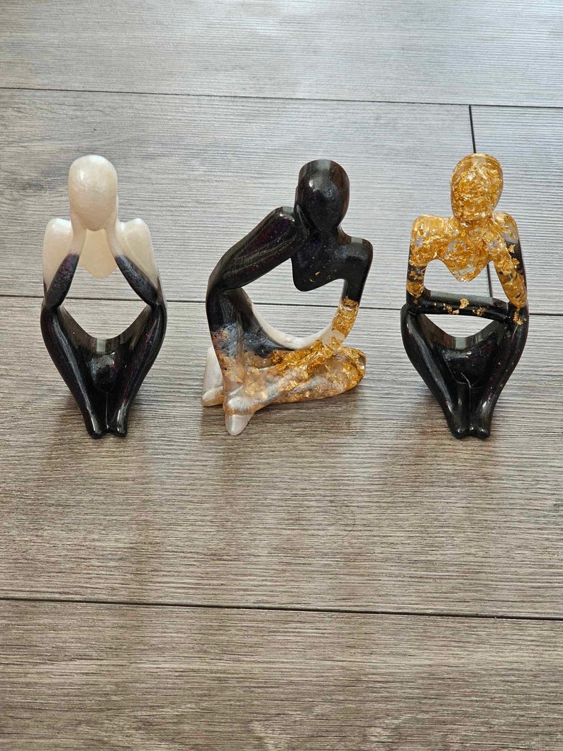 May include: Three abstract resin sculptures of people in a seated pose. The sculptures are painted in black, white, and gold.