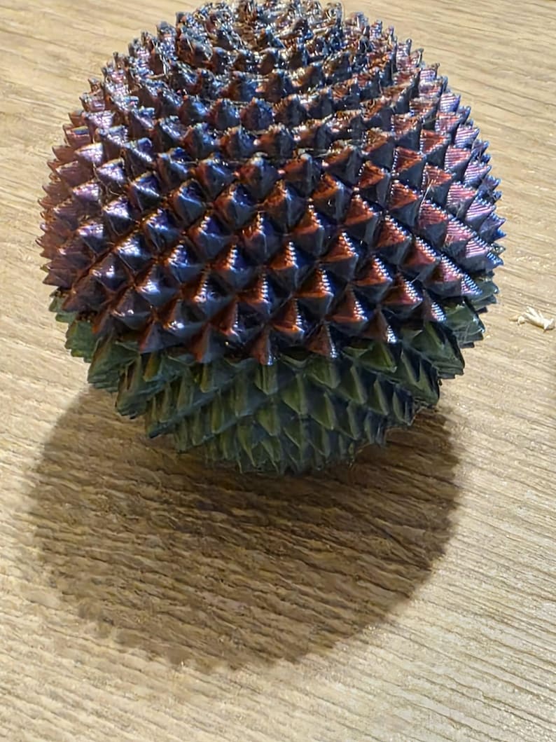 May include: A spherical object with a textured surface, featuring a pattern of small, pointed protrusions. The color transitions from a deep blue at the top to a green hue at the base, with hints of red and purple throughout.