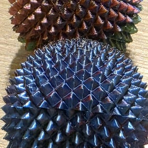 May include: Two spiky, spherical objects with a metallic sheen. The top one is a mix of brown, red, and green, while the bottom one is a deep blue. The surfaces are covered in sharp, triangular protrusions, creating a textured appearance.