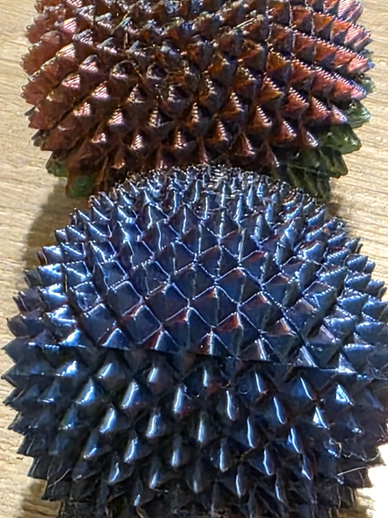 May include: Two spiky, spherical objects with a metallic sheen. The top one is a mix of brown, red, and green, while the bottom one is a deep blue. The surfaces are covered in sharp, triangular protrusions, creating a textured appearance.