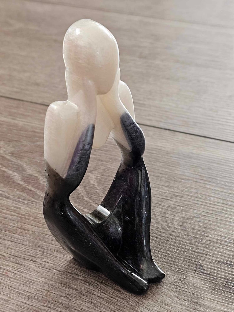 May include: A white and black abstract sculpture of a person sitting with their head in their hands.