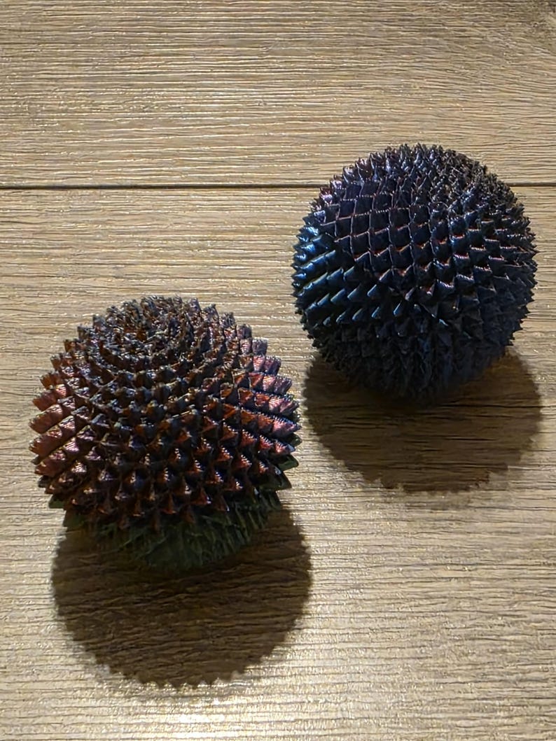 May include: Two spherical, spiky decorative objects. One is a mix of red, blue, and green hues, while the other is primarily blue with hints of purple. Both sit on a wooden surface, casting shadows.