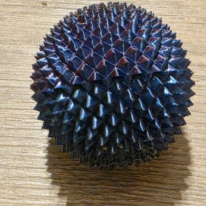 May include: A dark blue, spiky, spherical object with a textured surface. The object has a geometric pattern of triangular shapes and a glossy finish. The overall shape is round and symmetrical, with a slight shadow visible.
