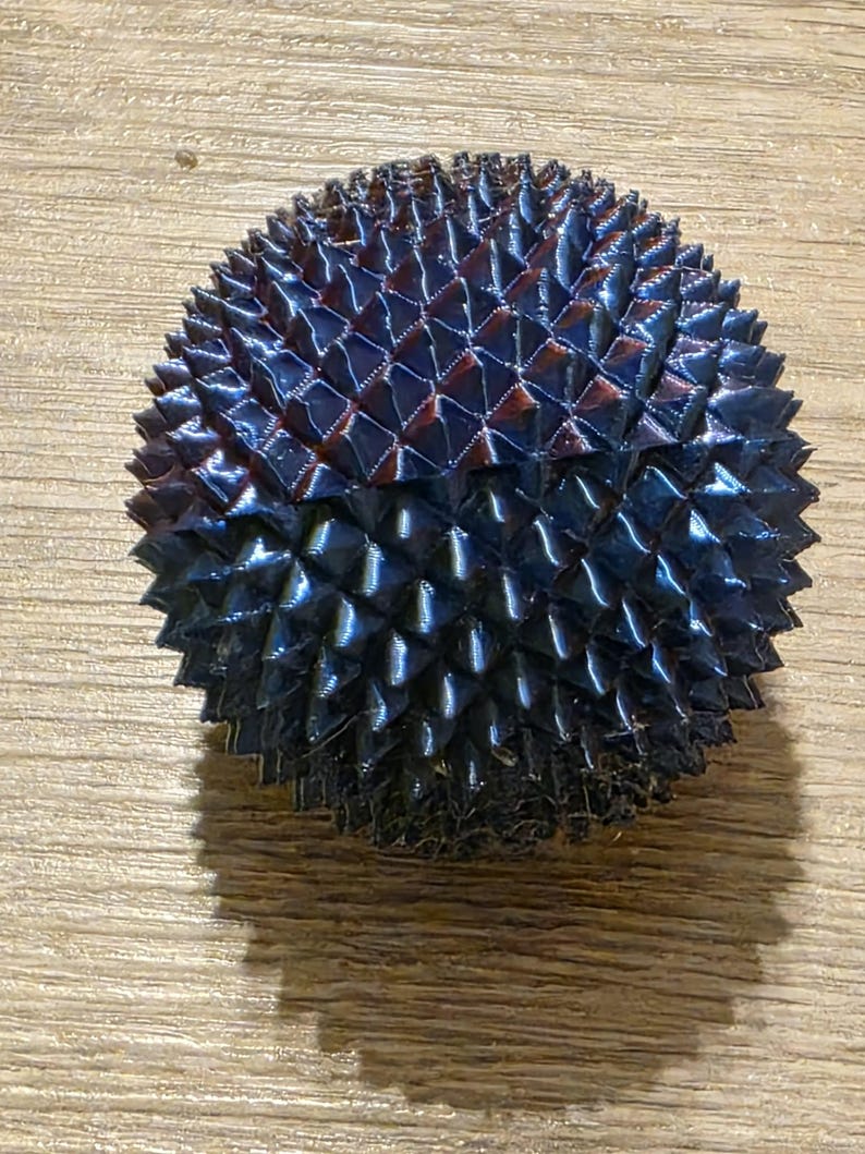 May include: A dark blue, spiky, spherical object with a textured surface. The object has a geometric pattern of triangular shapes and a glossy finish. The overall shape is round and symmetrical, with a slight shadow visible.