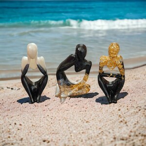 May include: Three small resin sculptures of people sitting with their heads down. The sculptures are painted black and white with gold accents. The sculptures are sitting on a sandy beach.