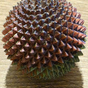 May include: A spherical, iridescent object with a textured surface. The object is covered in small, triangular protrusions, creating a spiky appearance. The colors shift from bronze to green and blue, giving it a unique, eye-catching look. The object is likely a decorative item.