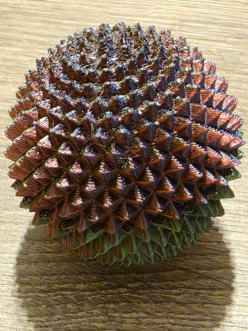May include: A spherical, iridescent object with a textured surface. The object is covered in small, triangular protrusions, creating a spiky appearance. The colors shift from bronze to green and blue, giving it a unique, eye-catching look. The object is likely a decorative item.