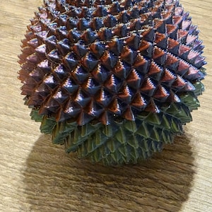 May include: A multi-colored, spiky sphere with a metallic sheen. The sphere transitions from green at the base to shades of blue, purple, and copper at the top. The object is resting on a wooden surface.