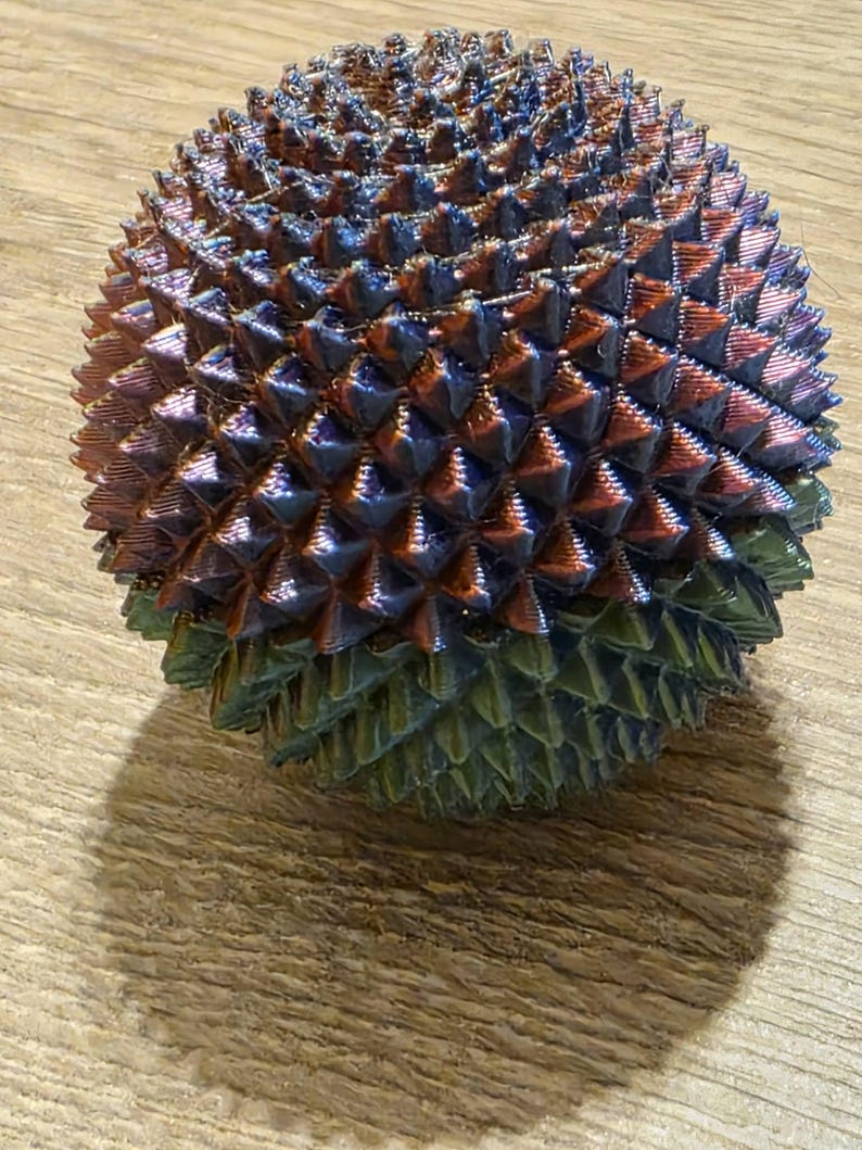 May include: A multi-colored, spiky sphere with a metallic sheen. The sphere transitions from green at the base to shades of blue, purple, and copper at the top. The object is resting on a wooden surface.