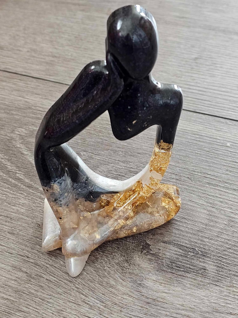 May include: A black and gold resin sculpture of a person sitting with their head resting on their hand. The sculpture has a modern abstract style.