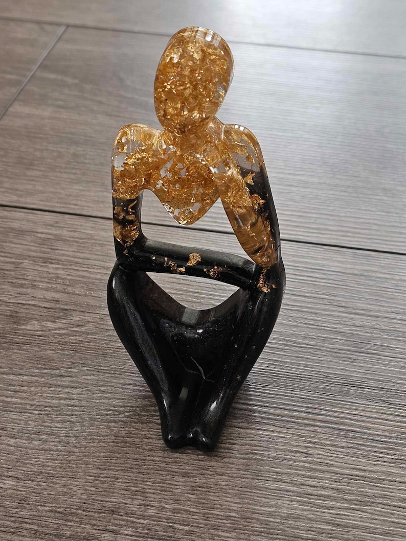 May include: A black and gold resin sculpture of a person sitting with their legs crossed. The sculpture is made of resin and has a gold leaf finish.