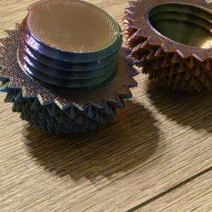 May include: Two iridescent, gear-shaped objects with a metallic sheen. One is fully assembled, showing a threaded base and a ridged top. The other is disassembled, revealing the interior. The objects sit on a wooden surface.