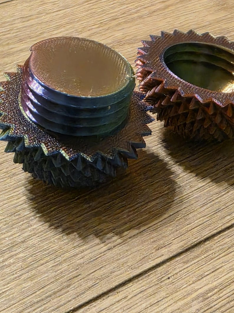 May include: Two iridescent, gear-shaped objects with a metallic sheen. One is fully assembled, showing a threaded base and a ridged top. The other is disassembled, revealing the interior. The objects sit on a wooden surface.