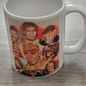 May include: White ceramic mug with a collage of professional wrestlers. The mug has a large handle and a colourful design with multiple faces, including the text "HOGAN" on a headband.