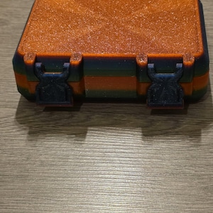 May include: A small, rectangular storage box with an orange glittery top and a multi-coloured body. The box has two black clasps on the front and is likely made of plastic. The box is closed and sits on a wooden surface.