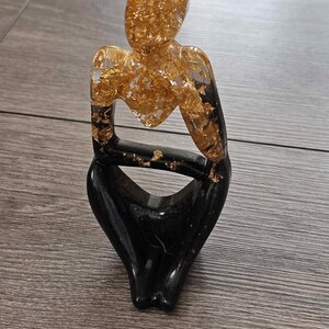 May include: A black and gold resin sculpture of a person sitting with their legs crossed. The sculpture is made of clear resin with gold leaf flakes embedded in it.