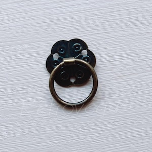 May include: A black metal ring pull handle with a decorative floral design. The handle is attached to a white surface.