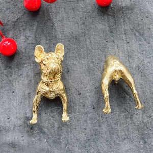 May include: Two gold-coloured metal figurines of dogs. One is standing with its head up and the other is sitting with its head down. The figurines are on a grey fabric background.