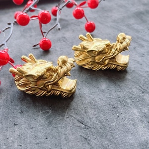 May include: Two gold dragon head figurines with intricate details. The figurines are on a gray textured surface.