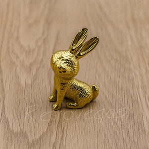 May include: A gold-coloured rabbit figurine with a textured surface. The rabbit is sitting on a wooden surface.