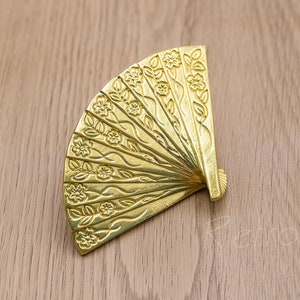 May include: Two gold-coloured fan-shaped cabinet pulls with an embossed floral pattern. They are set against a light wood grain surface. These decorative hardware pieces add a touch of elegance to any furniture.