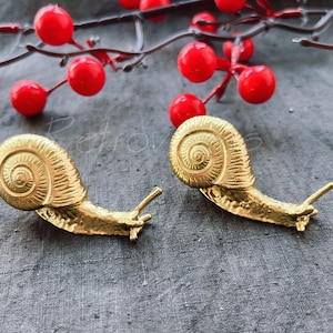 Peut inclure: Deux sculptures d'escargots en or avec des motifs de coquille détaillés. Les escargots sont placés sur un fond de tissu gris.