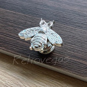 Könnte beinhalten: Ein silberfarbener, bienenförmiger dekorativer Ansteckpin. Die Biene hat detaillierte Flügel und Körpersegmente. Der Pin befindet sich auf einer dunklen Holzmaserungsoberfläche. Das Wort "Retrovegaz" steht unten in Schreibschrift.