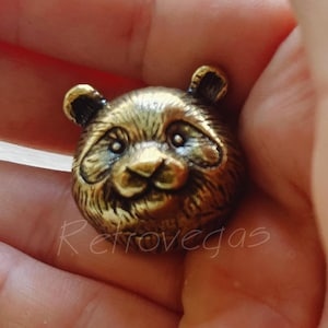 May include: A detailed, antique-style panda bear head figurine in a bronze finish. The panda has a smiling expression, with visible eyes, nose, and ears. The figurine is held in a hand.