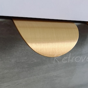 May include: Close-up of two gold-coloured, teardrop-shaped cabinet pulls. The pulls are mounted on a grey, textured surface. The word "Retrovegas" is faintly visible in the lower left corner.