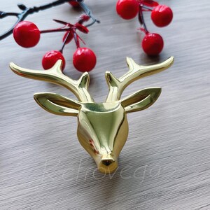 Op de afbeelding: Een gouden rendierkop ornament met een eenvoudig ontwerp. Het ornament is gemaakt van metaal en heeft een gladde, glanzende afwerking.