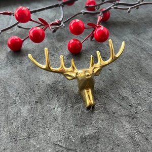 Könnte beinhalten: Ein goldfarbener Hirschkopf-Wandhaken mit Geweih. Der Haken befindet sich auf einer grauen, strukturierten Oberfläche.