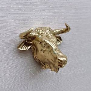 May include: A gold-coloured bull's head sculpture with detailed features. The sculpture is mounted on a white wall.