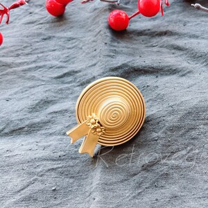 Könnte beinhalten: Eine goldfarbene Hutnadel mit einem Strohhut-Design und einer kleinen Schleife. Die Nadel befindet sich auf einem grauen Stoffhintergrund.