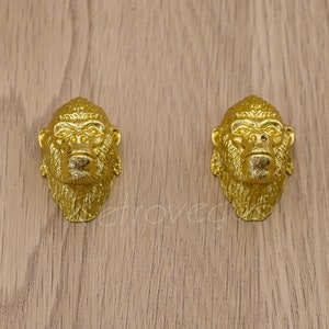 Puede incluir: Dos figuritas de cabeza de gorila de color dorado. Las cabezas de gorila están mirando hacia adelante con la boca cerrada. Las figuritas están sobre una superficie de madera marrón claro.