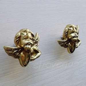 Könnte beinhalten: Zwei goldene Schrankknöpfe in Form von Cherubim mit Flügeln und einem lächelnden Gesicht. Die Knöpfe befinden sich auf einem weißen Hintergrund.