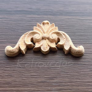 May include: A light-colored, wooden decorative architectural element with a symmetrical design. The piece features a central floral motif with curved, scroll-like details on either side. The item is set against a wood-grain background.