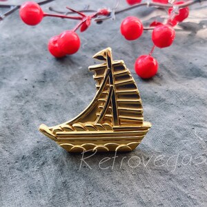May include: A gold-coloured metal sailboat drawer pull with a detailed design. The pull is on a grey fabric surface.