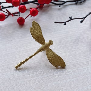 May include: A gold dragonfly brooch with detailed wing patterns. The brooch is set against a white wooden background, with red berries and branches in the background. The word "Retrovegas" is faintly visible.