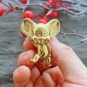 May include: A gold-coloured figurine of a cartoon mouse with large ears and a wide smile. The figurine is standing on a hand.