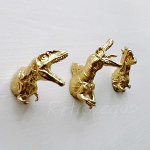 Puede incluir: Tres ganchos de pared de dinosaurio de color dorado. Los dinosaurios están en una variedad de poses, con la boca abierta y las garras extendidas.