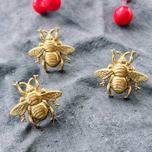 Könnte beinhalten: Drei goldene Broschen in Bienenform auf einem grauen Stoffhintergrund. Die Bienen haben detaillierte Flügel und Körper.