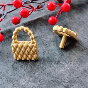 May include: Two gold-toned cabinet knobs. One is shaped like a small handbag with a quilted design. The other is a simple round knob with a decorative edge.
