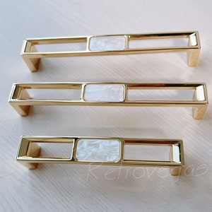 May include: Three gold cabinet pulls with a white rectangular insert. The pulls have a simple, modern design.