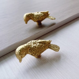 May include: Two gold bird-shaped drawer pulls. The pulls are detailed with a textured finish and have a slightly curved shape.